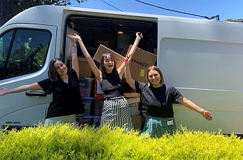 Three femal graduates raising their hands and similing ion the sides of a van with its door open.