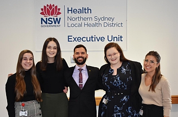 One male and four female graduates standing and similing in front of a sign that reads NSLHD Executive Unit